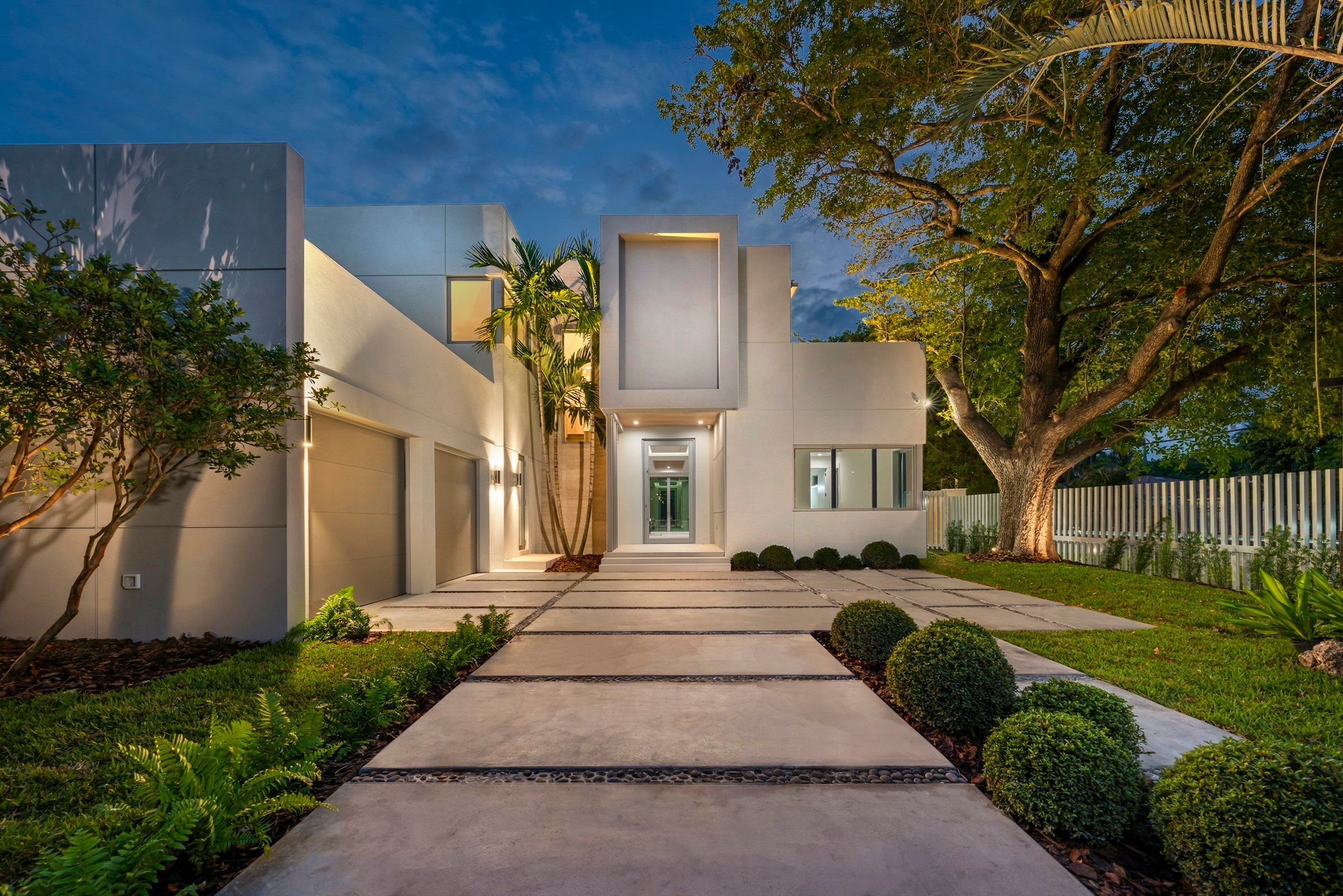 A lit pathway between clipped hedges leads to a modern house at dusk